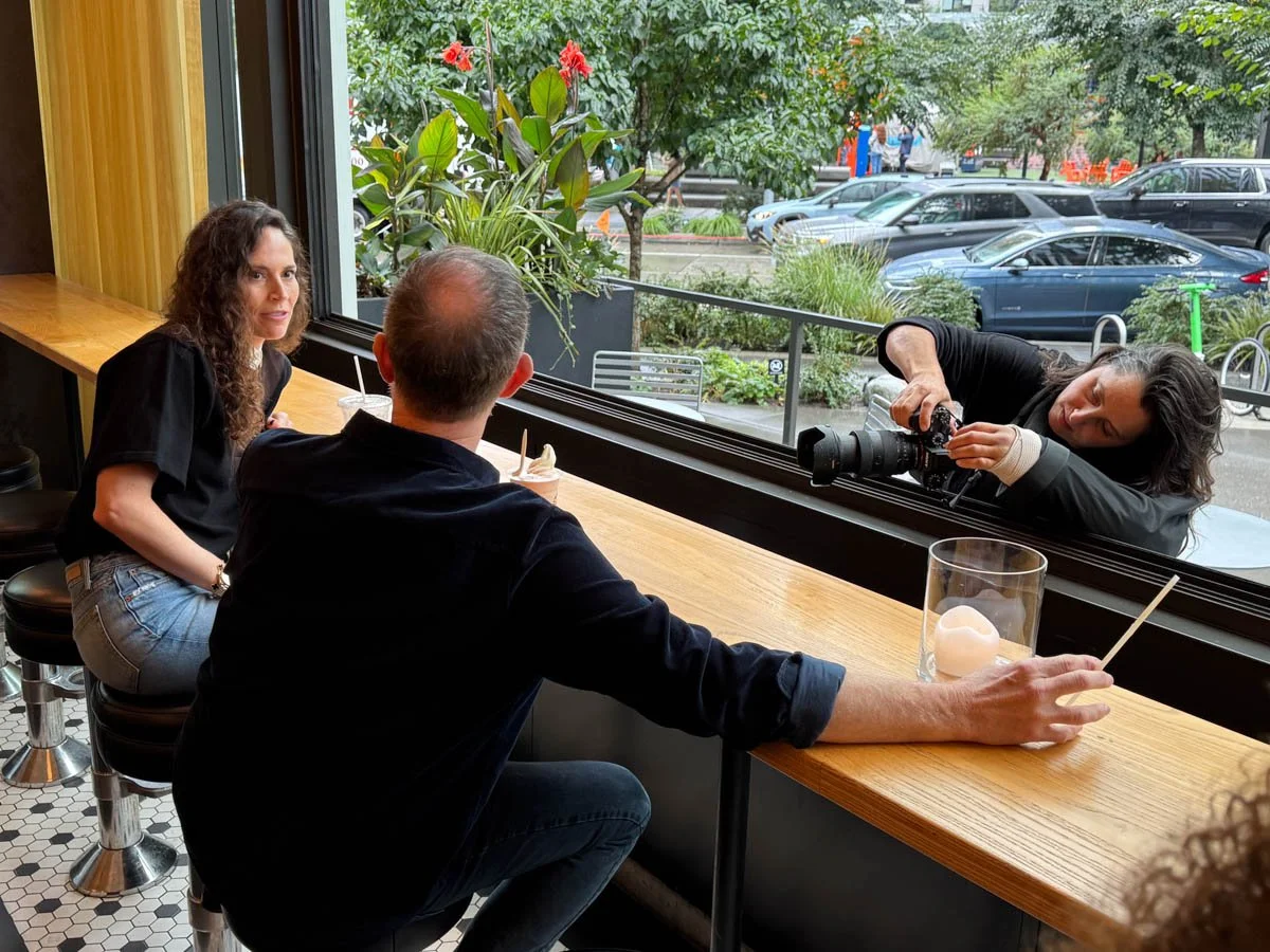 A woman is taking a photograph of two people sitting at a cafe table near a window, with cars and trees outside.