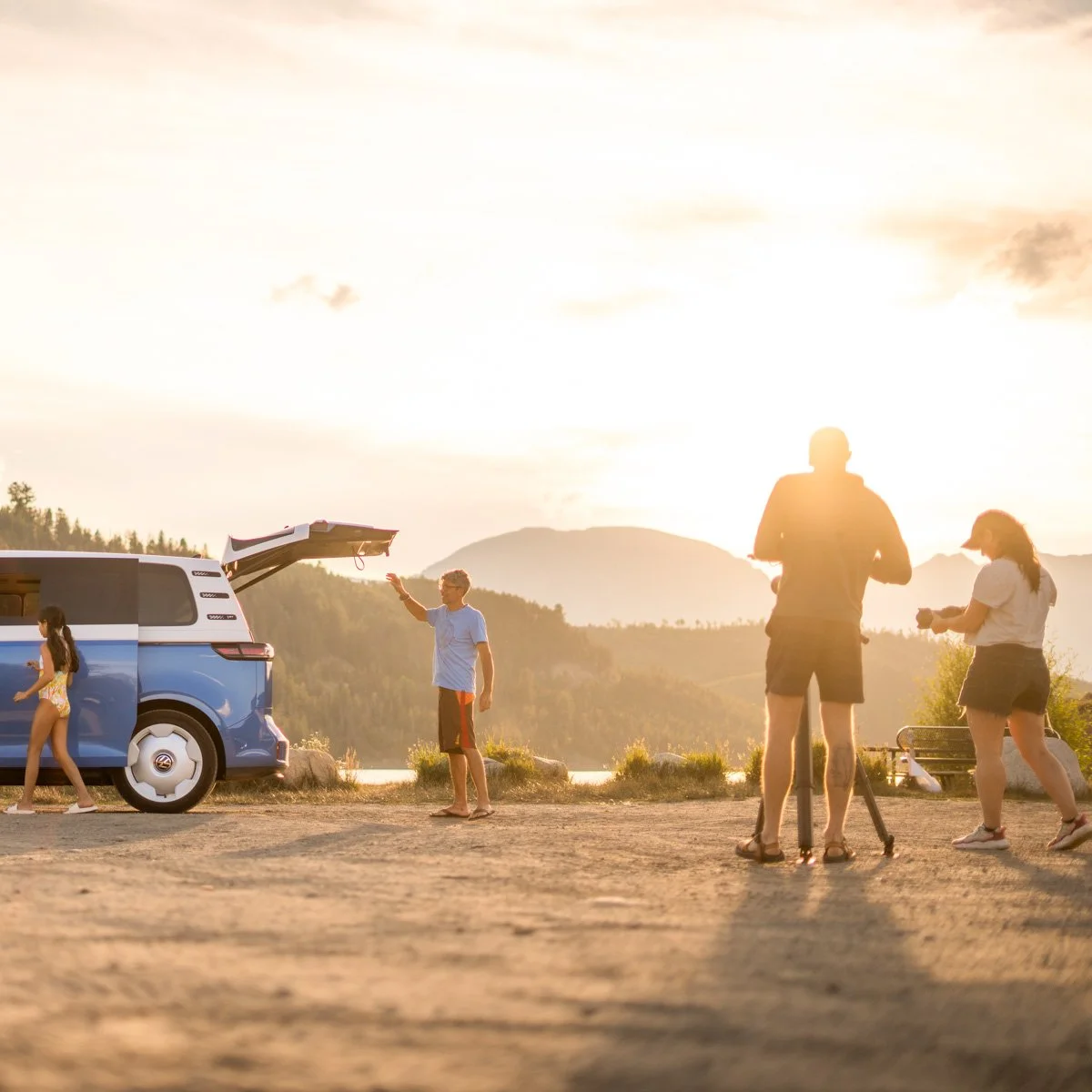 A group of people enjoying a sunset near a lake, with a man waving at a blue van and a girl walking towards it, while others take photos in front of scenic mountains.