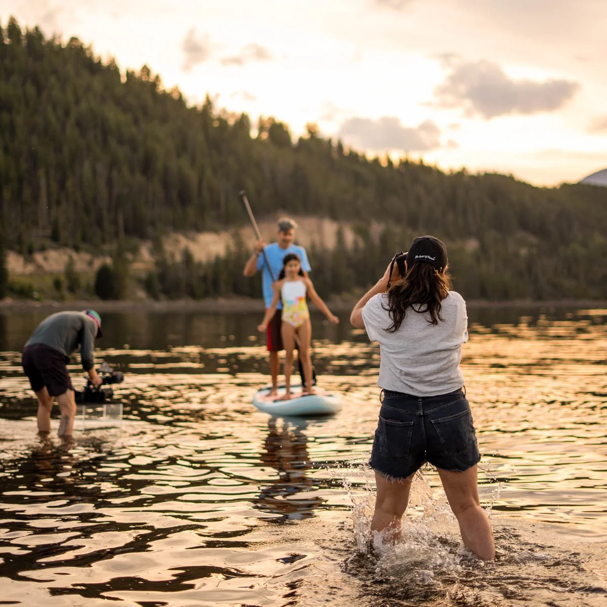 A woman takes a photo of two children on a paddleboard in a lake during sunset, with other people preparing to film and take pictures nearby.