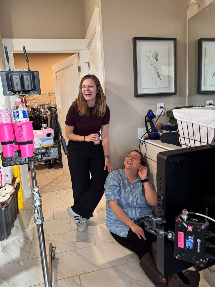 Two women on a film set, one standing and laughing, the other sitting on the floor, also laughing, in a room with equipment and decor.