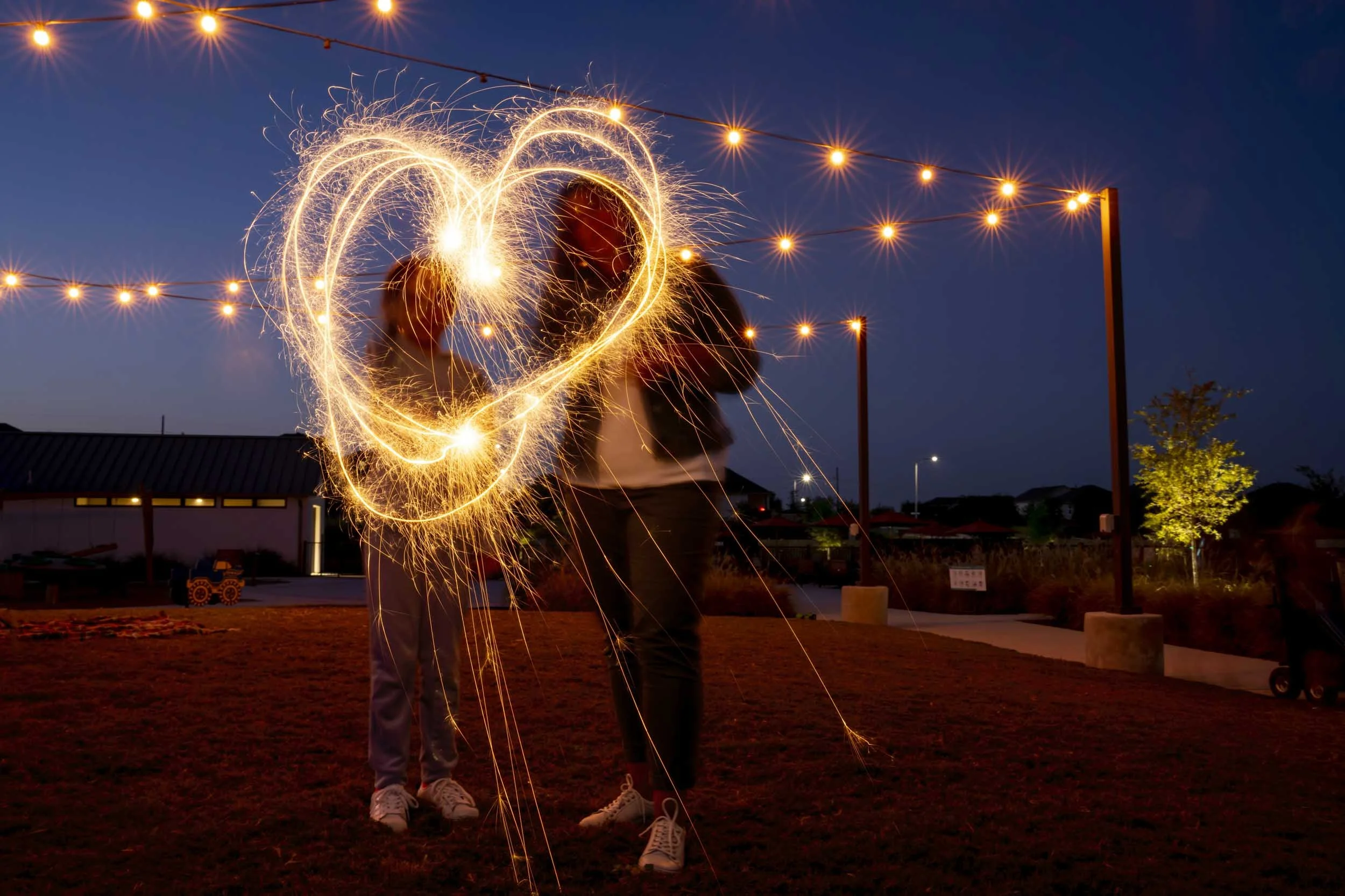 Two people draw a shape with sparklers at night, creating light trails and a heart in the air, with string lights overhead and trees in the background.
