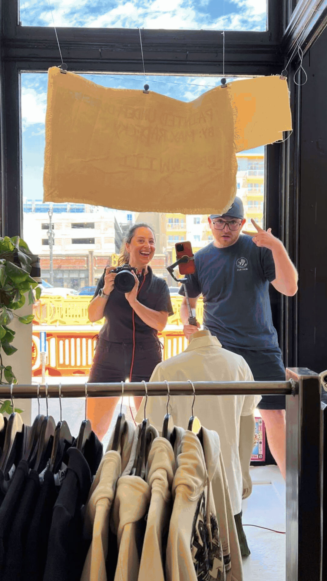 Two people taking a mirror selfie inside a clothing store, with racks of clothes in the foreground and a large window with a sunny sky in the background.