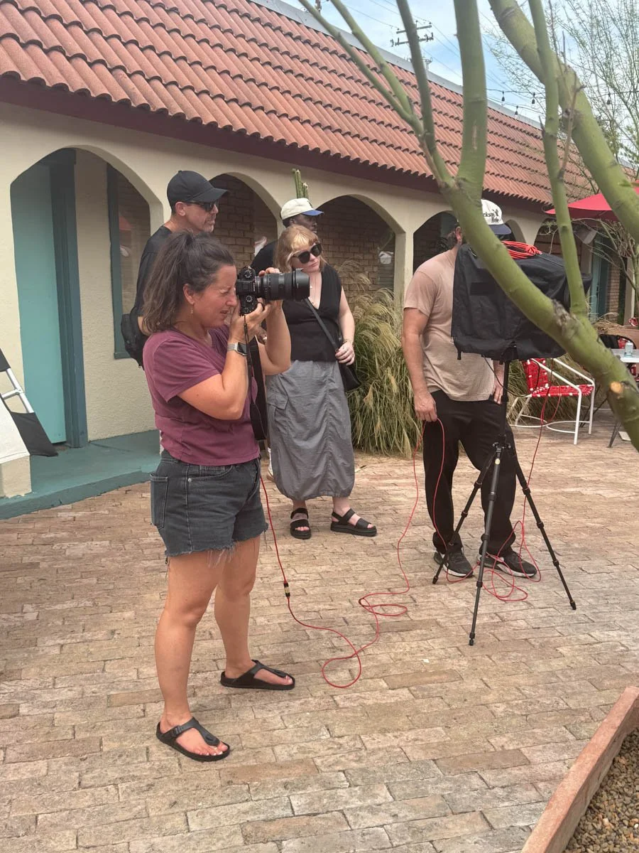 Group of people filming outdoors on a brick patio, with a woman holding a camera and other crew members around her, in front of a building with arched doorways and a red tile roof.