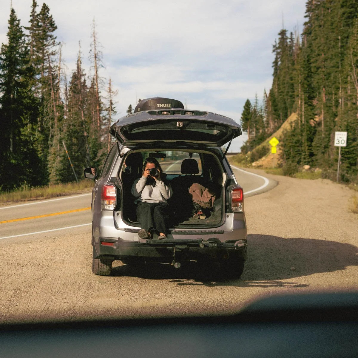 Person sitting in the open trunk of a car taking a photo, parked beside a winding mountain road surrounded by trees.