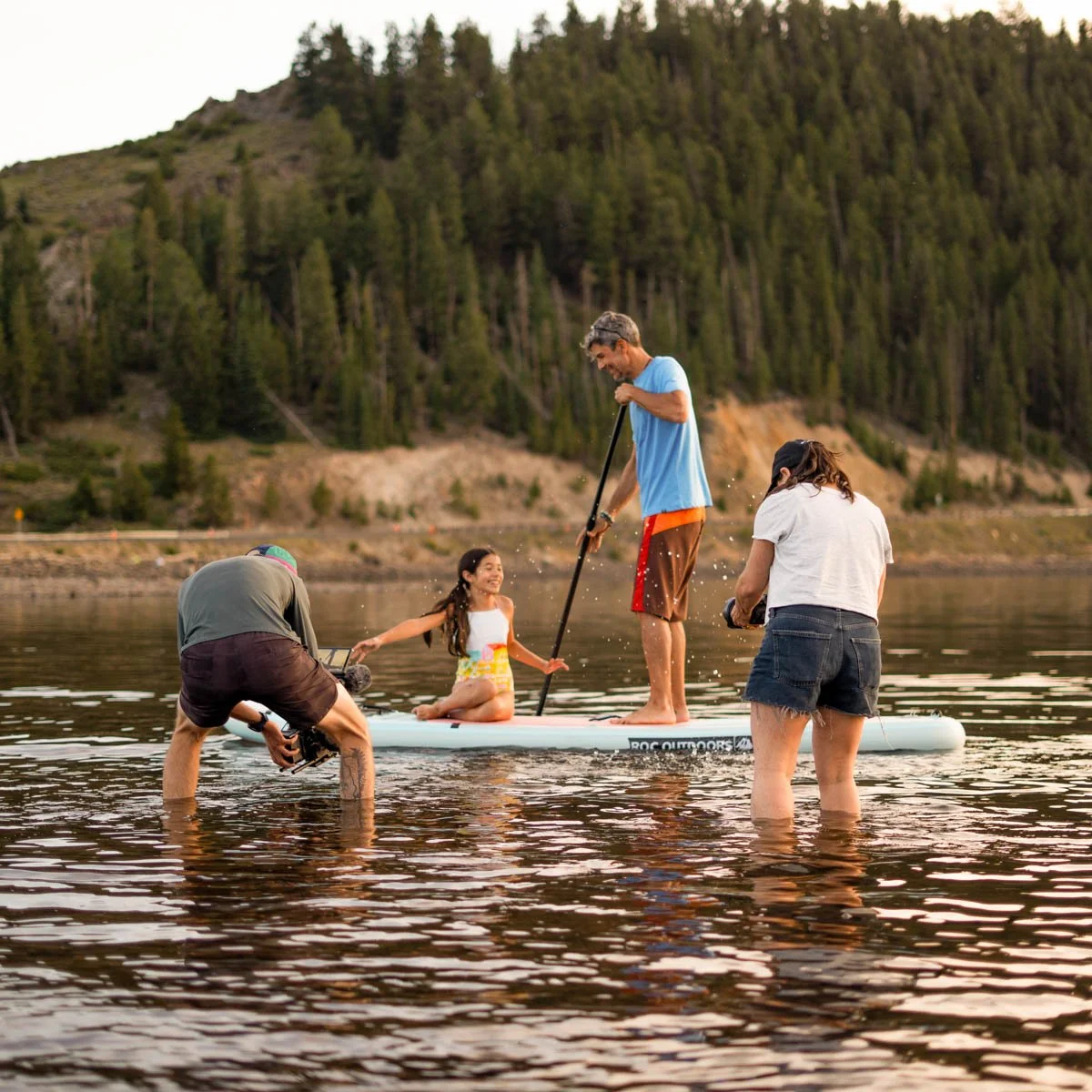 A family enjoying time on a stand-up paddleboard on a calm river with a forested mountain in the background.