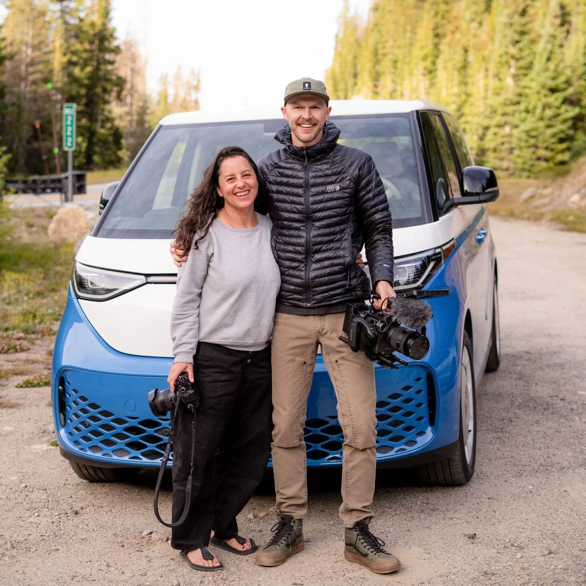 Two people, a woman and a man, standing in front of a blue and white electric vehicle in a forested area, smiling, with the woman holding a camera and the man holding a professional video camera.