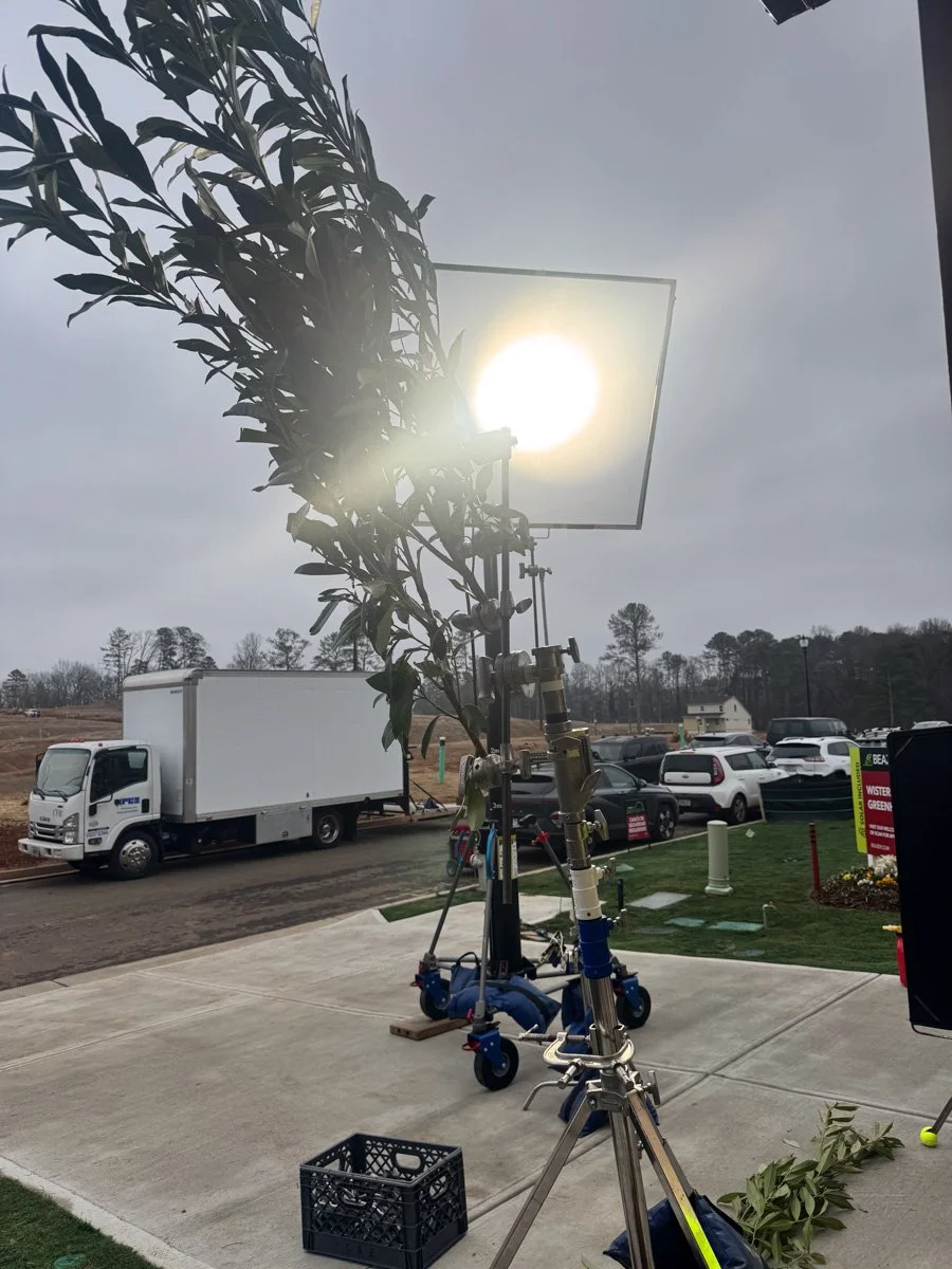Cinematic camera setup on a dolly with a matte box and a large filter, aimed at the sun, outdoors in a parking lot with parked cars and trees in the background.