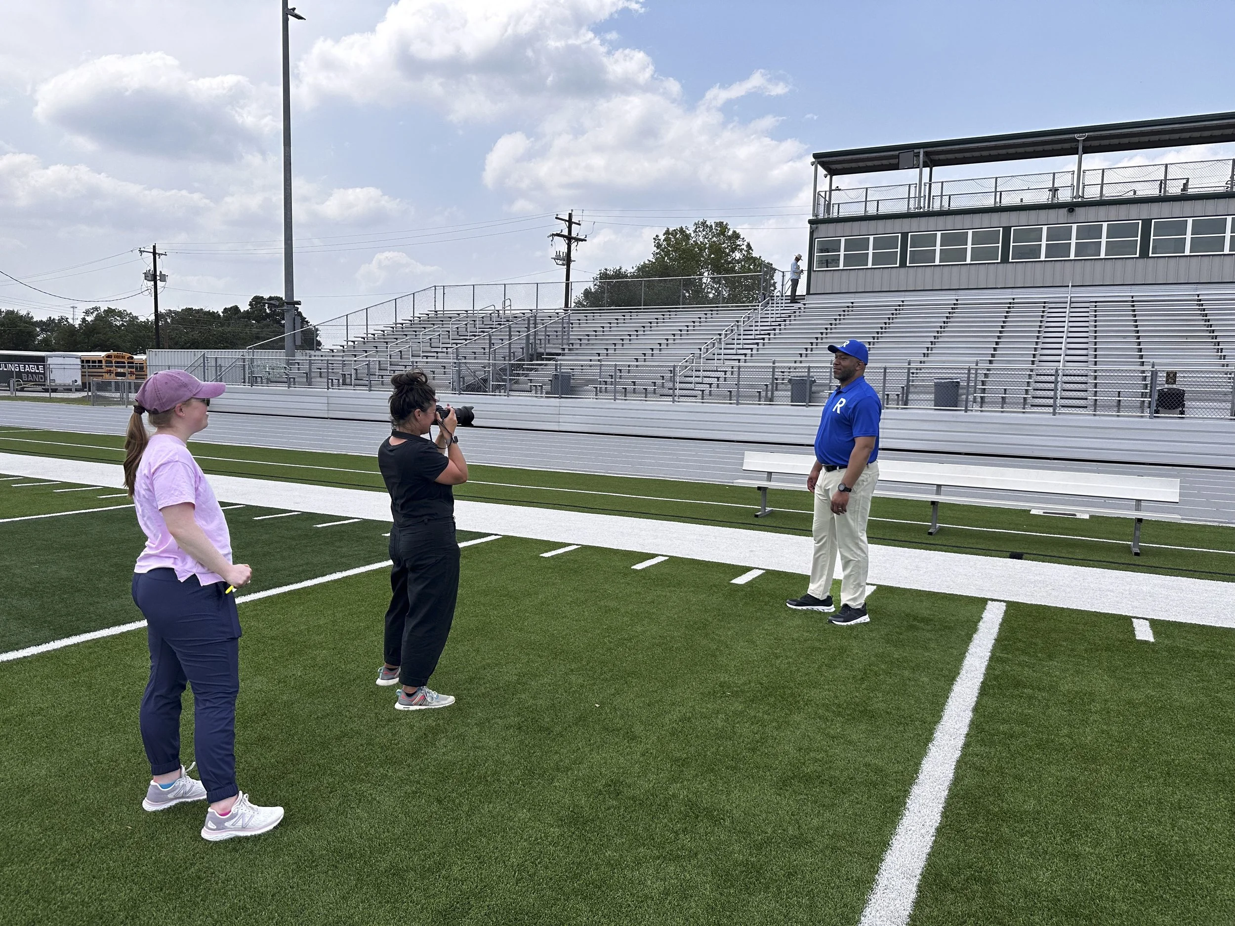 Three women on a football field with one taking a photograph of a man in a blue shirt and beige pants, standing near white yard line markings and empty bleachers in the background.