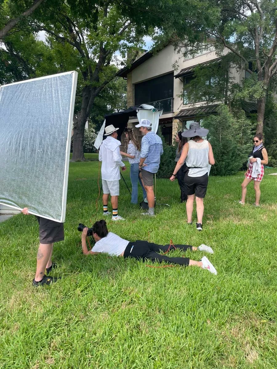 Filming crew on a grassy lawn filming scenes with a group of people at a house, with a large reflector and a woman lying on the grass taking photographs