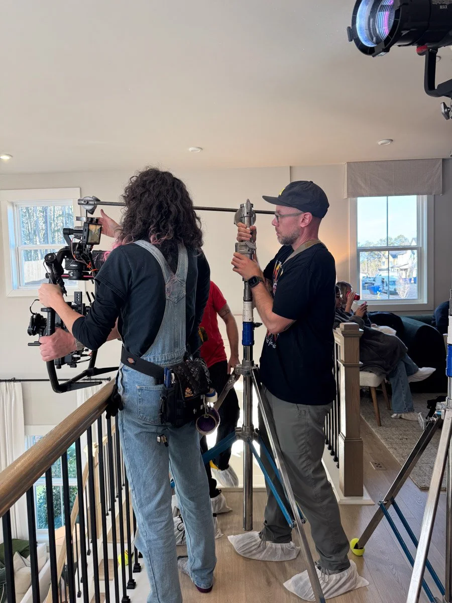 Two filmmakers, a woman with curly hair wearing overalls and a man with a cap, are filming inside a house. The woman is operating a camera mounted on a stabilizer, and the man is handling a tall lighting pole. They are standing on a Land without shoe