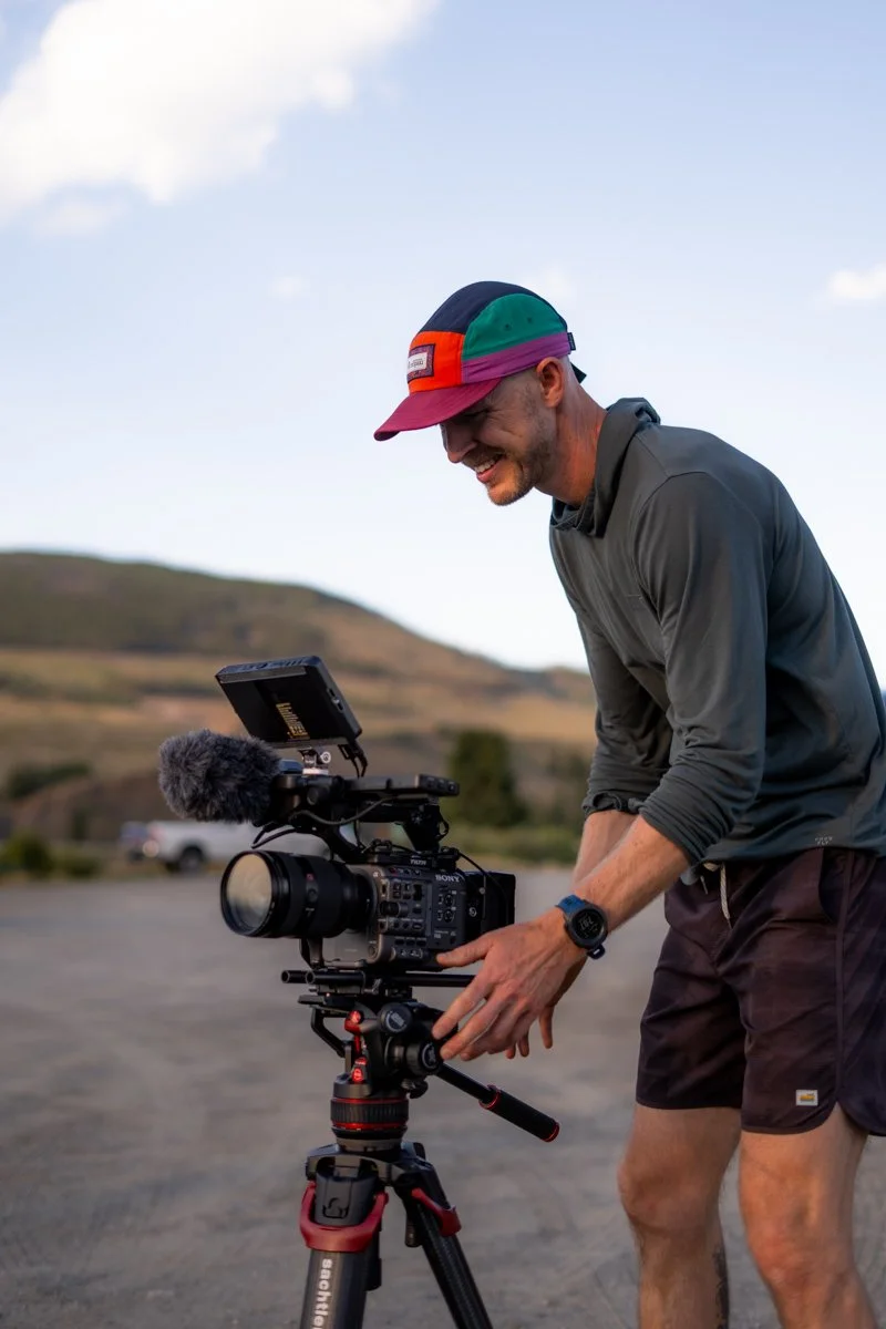 A man is setting up a video camera outdoors on a tripod during daytime, with rolling hills and a cloudy sky in the background.