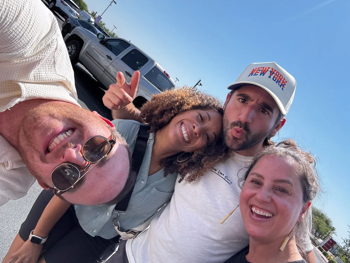 Group of four people taking a selfie outdoors on a sunny day, with parking lot and cars in the background.