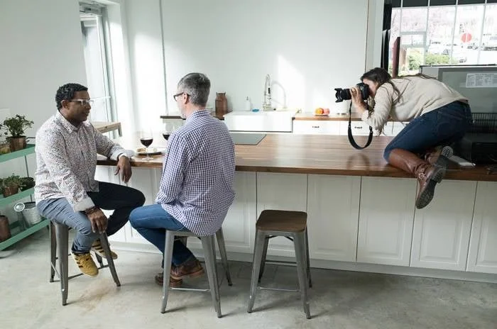 Two men sitting at a kitchen island, facing each other, while a woman takes their photograph from a countertop.