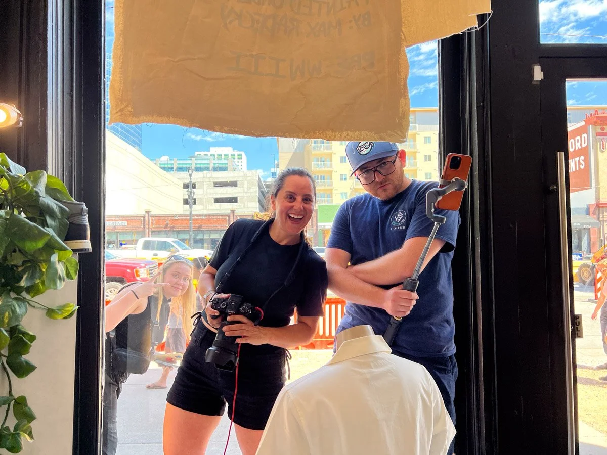 Two people taking a selfie inside a shop with a mannequin. Outside, a woman is making a peace sign and smiling. City buildings and cars are visible through the window.