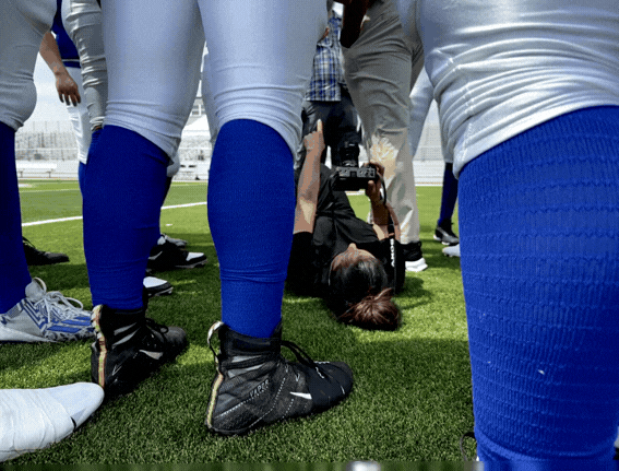 A woman lying on her back taking a selfie at a sports field surrounded by people in athletic clothing.