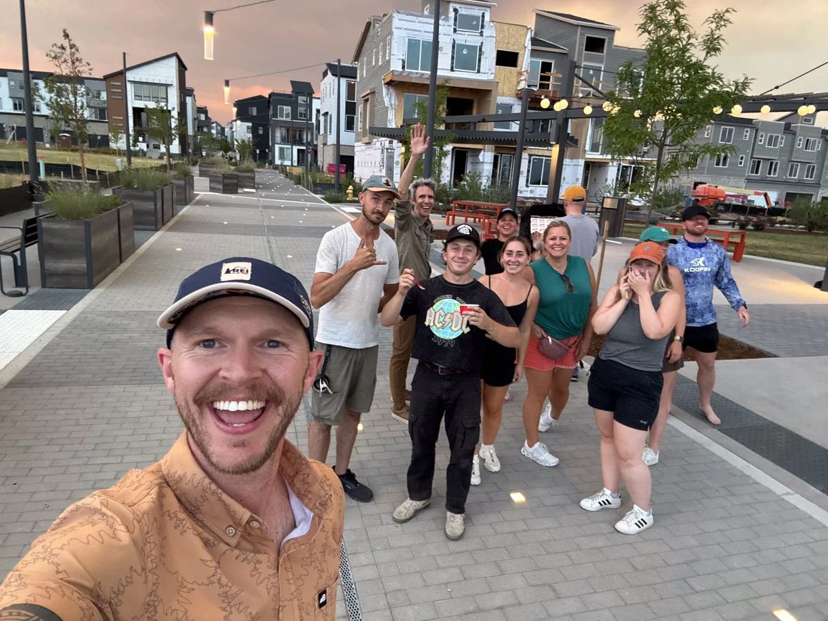 Group of people smiling and posing for a selfie on a modern, outdoor urban walkway during sunset, with new construction buildings in the background.