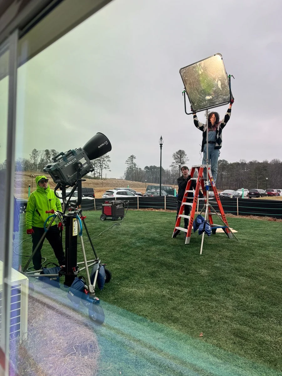 A woman standing on a ladder holding a large lighting reflector during a video shoot or photo shoot, with a man beside her and a crew member operating a photography light, outdoors on a grassy area with parked cars and trees in the background.