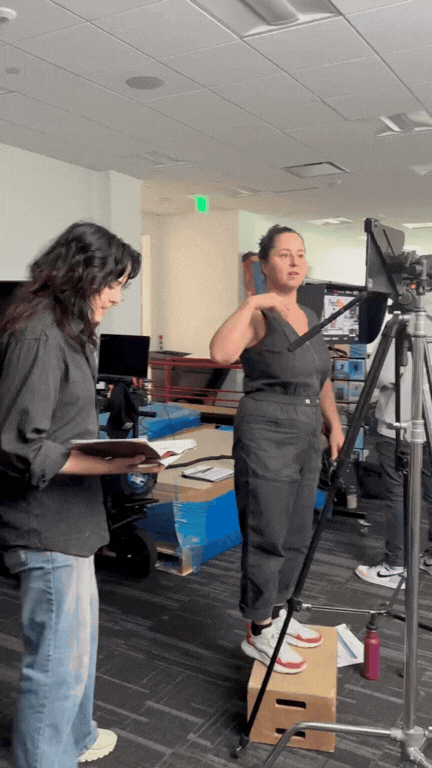 Two women working in a studio with camera equipment. One woman is adjusting a camera, while the other reads from a notebook.
