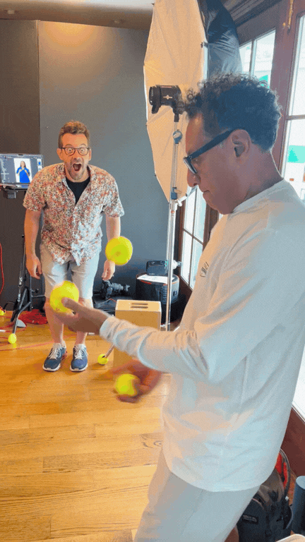 Two men indoors, one is juggling yellow balls while the other reacts with surprise.