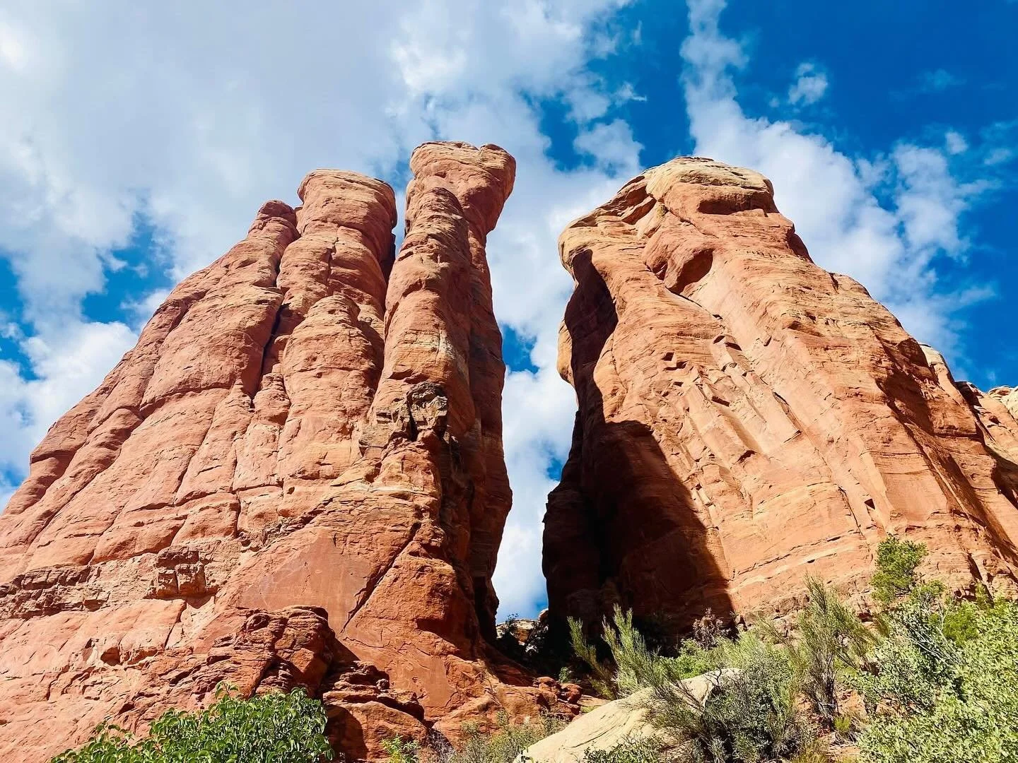 Tall red rock formations against a blue sky with scattered clouds and some green vegetation in the foreground.