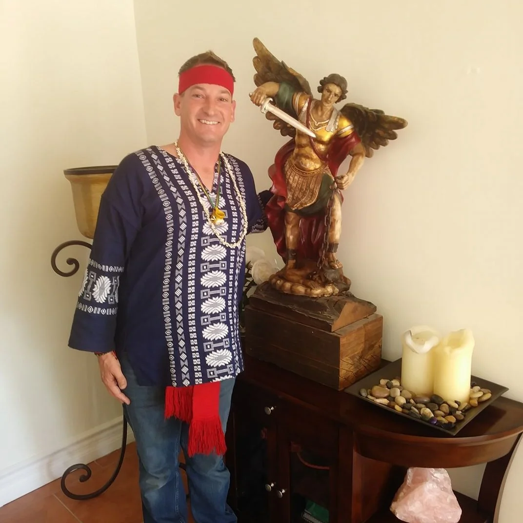 Man dressed in ethnically inspired clothing, wearing a red headband and red sash, standing next to a sculpture of an angel with wings, holding a sword, on a wooden table with candles and decorative stones.