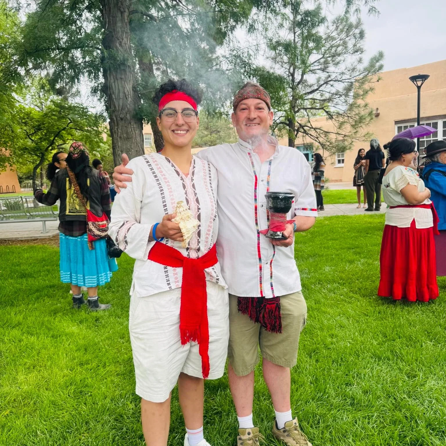 Two people dressed in traditional attire smiling at an outdoor event, with multiple other individuals in traditional clothing in the background.
