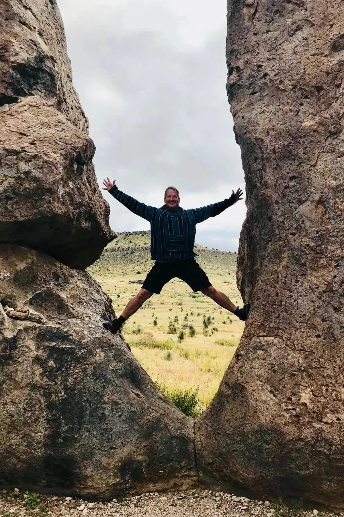 A man in a striped hoodie and shorts standing between two large rocks with arms and legs spread wide, smiling, in a desert landscape with grass and hills under an overcast sky.