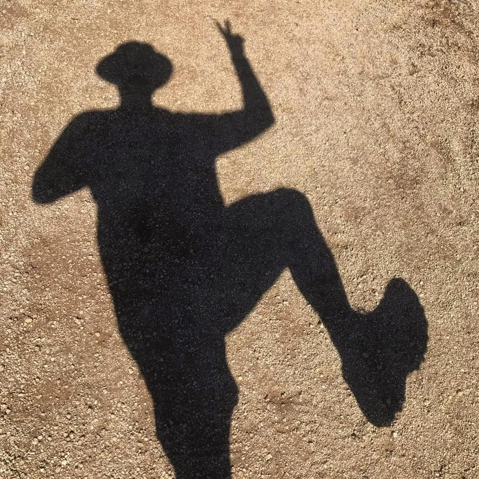 Shadow of a person on a gravel surface, making a peace sign with one hand and holding an object in the other.