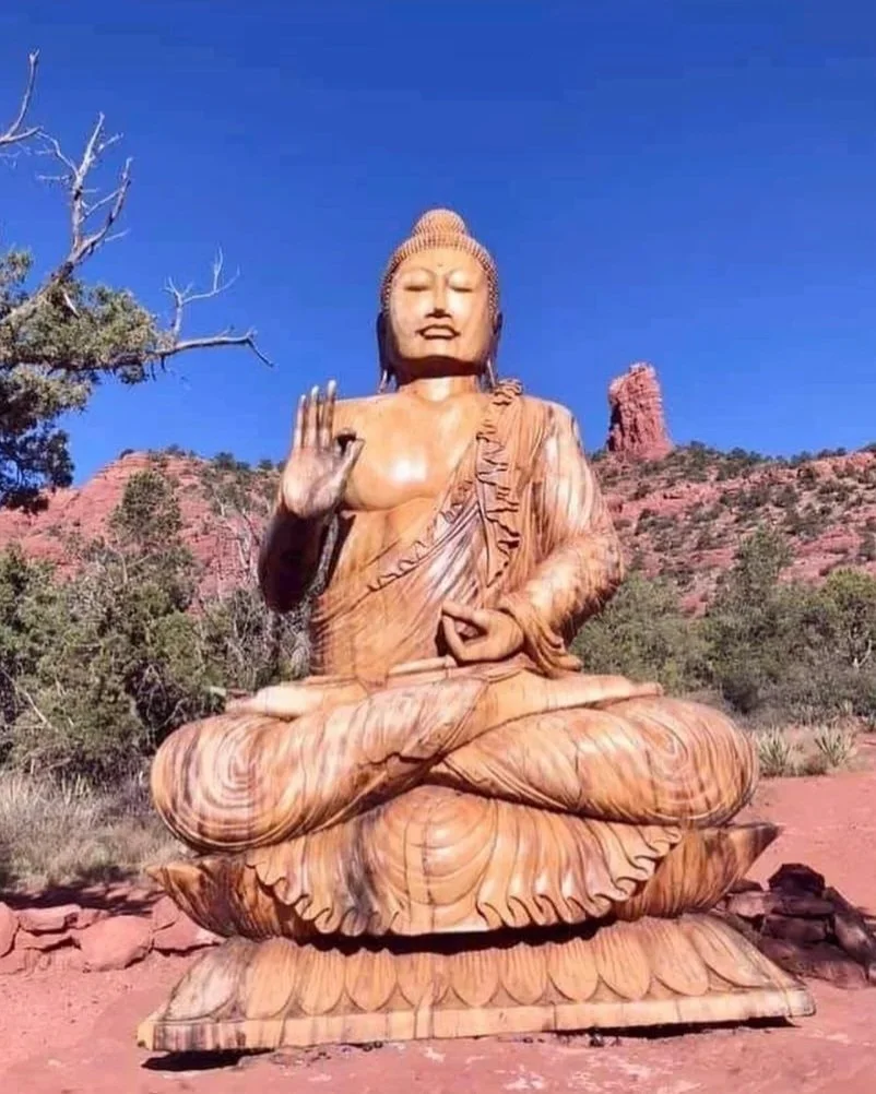 A large wooden sculpture of Buddha sitting cross-legged outdoors under a clear blue sky, with one hand raised in a gesture of blessing.