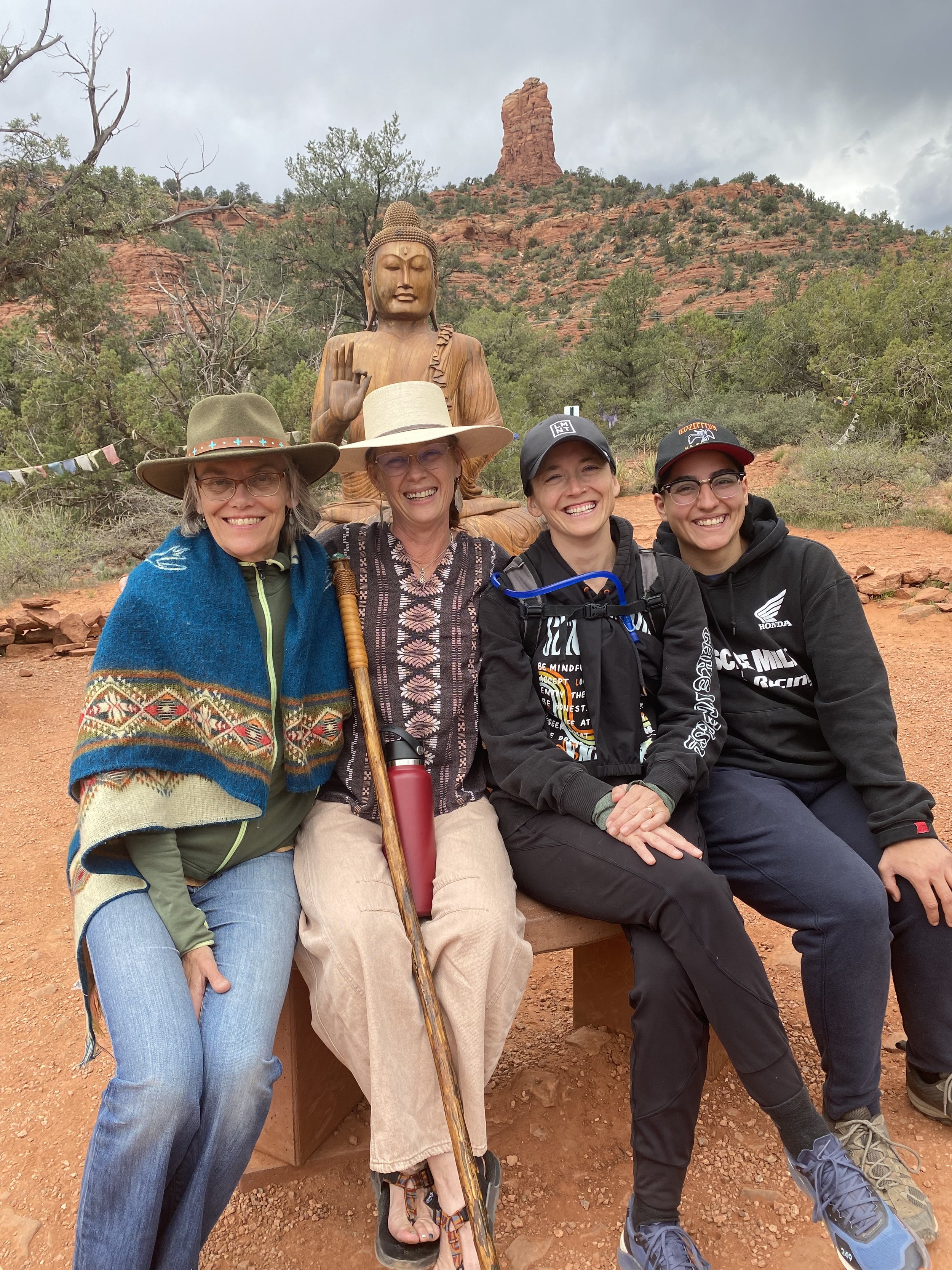 Four women sitting on a bench in front of a Buddha statue and a red rock formation in a desert landscape, with cloudy sky.