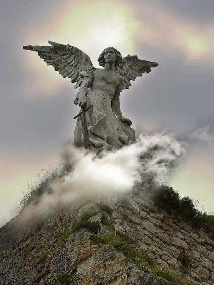 Statue of a winged female figure holding a sword, situated on a rocky hillside with clouds surrounding its base.
