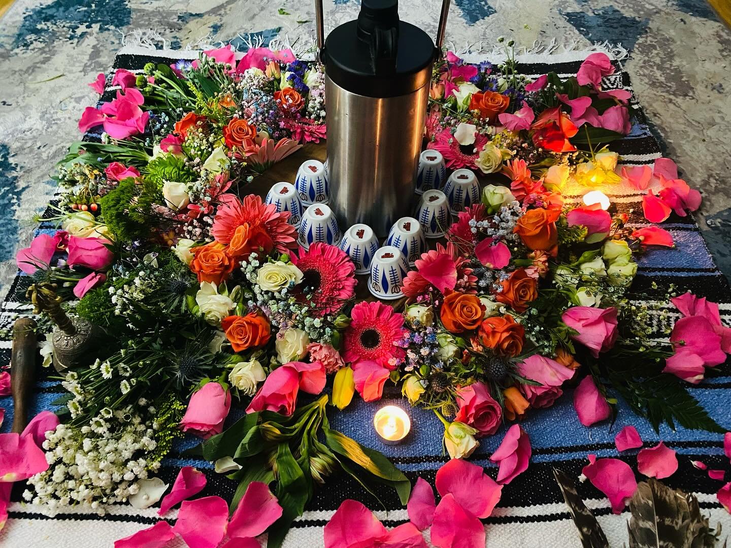 A flower arrangement with pink, orange, white, and purple flowers surrounding a stainless steel candle holder with a lit candle, placed on a striped black and white textile on a concrete surface.