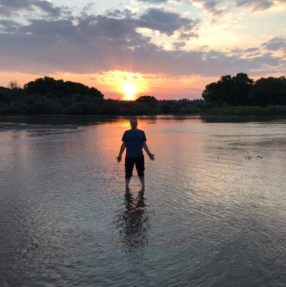 Person standing in shallow water at sunset with trees and clouds in the sky.