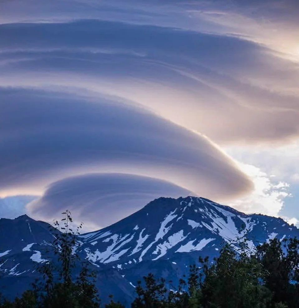 Mountain with snow patches under a sky with large, layered, swirling clouds during sunset.