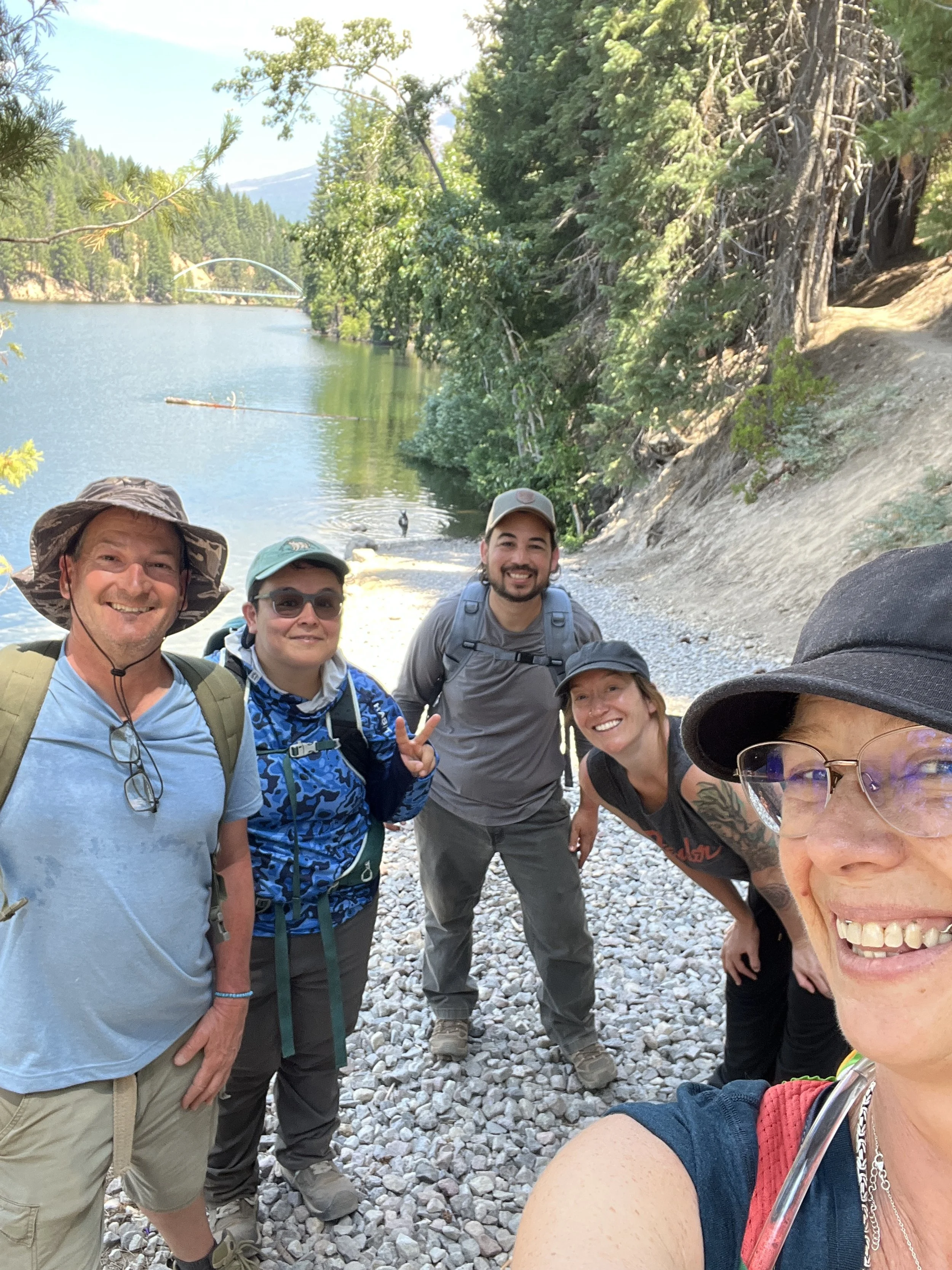 A group of five smiling hikers taking a selfie on a forested lakeside trail.