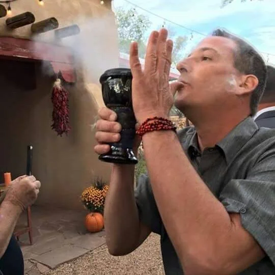 A man is outdoors, holding a black goblet to his lips as smoke rises from it, resembling smoking from a pipe or hookah. In the background, there are pumpkins and autumn decorations, indicating a fall setting.