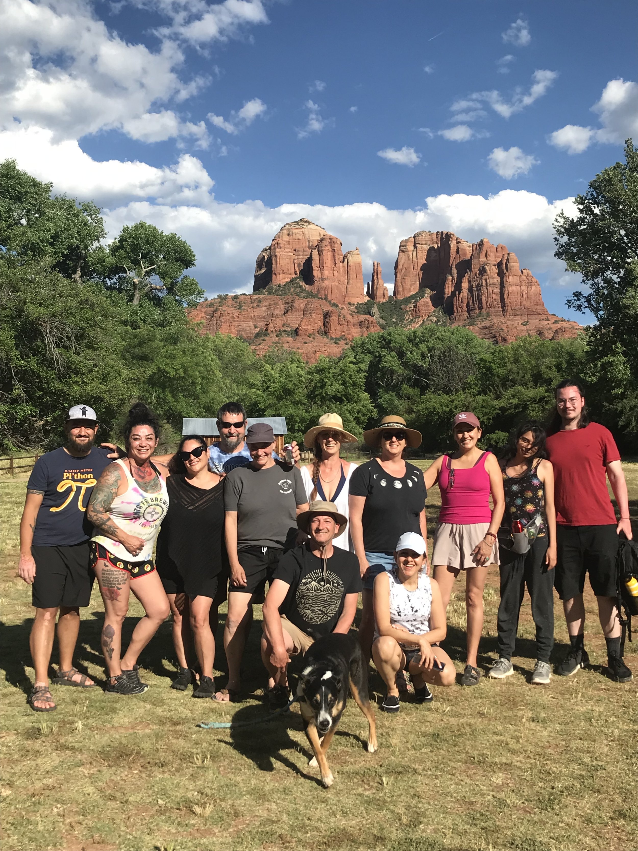 Group of people posing outdoors with red rock formations and green trees in the background, clear blue sky with some clouds.