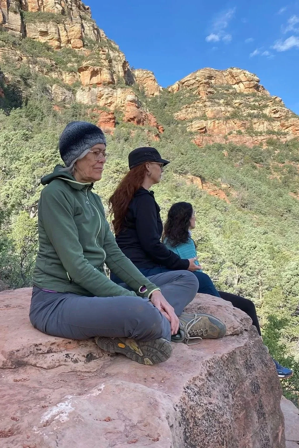 Three women sitting on a large rock outdoors with a mountainous background, green trees, and a blue sky.