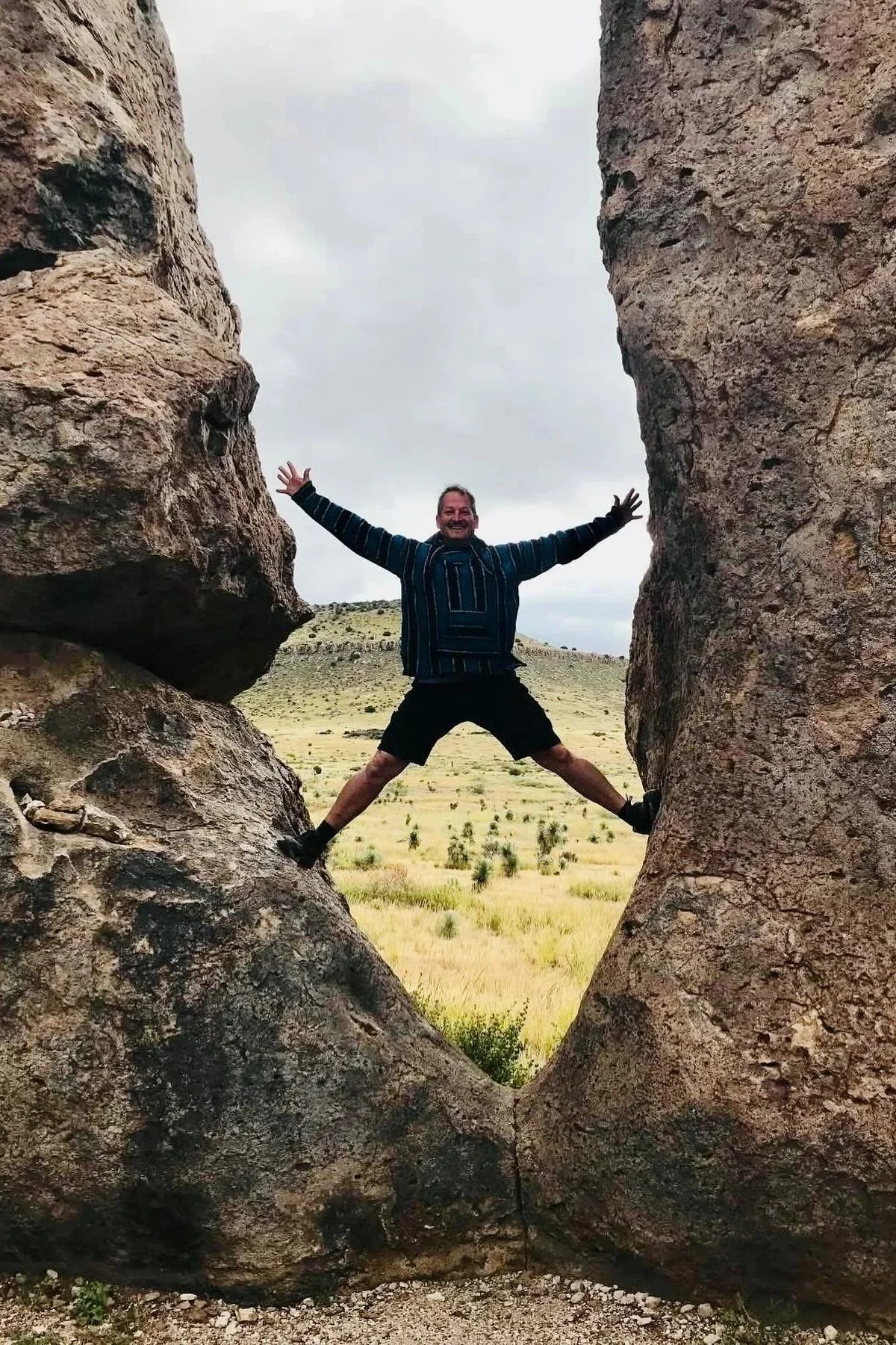 A man wearing a striped jacket and black shorts is standing between two large rocks with his arms and legs spread wide, creating the illusion of a star shape. The background shows a grassy plain with small bushes and a hill under a cloudy sky.