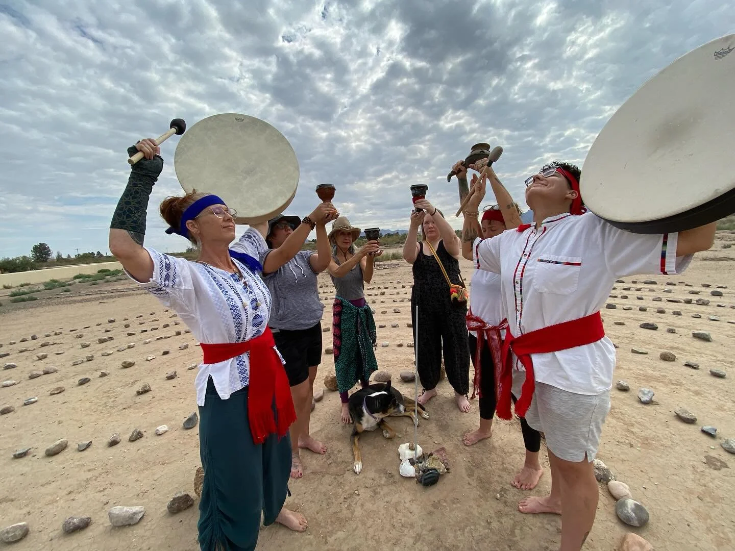 Group of people dressed in traditional clothing performing a ceremony with drums and chalices on a desert landscape with cloudy sky.