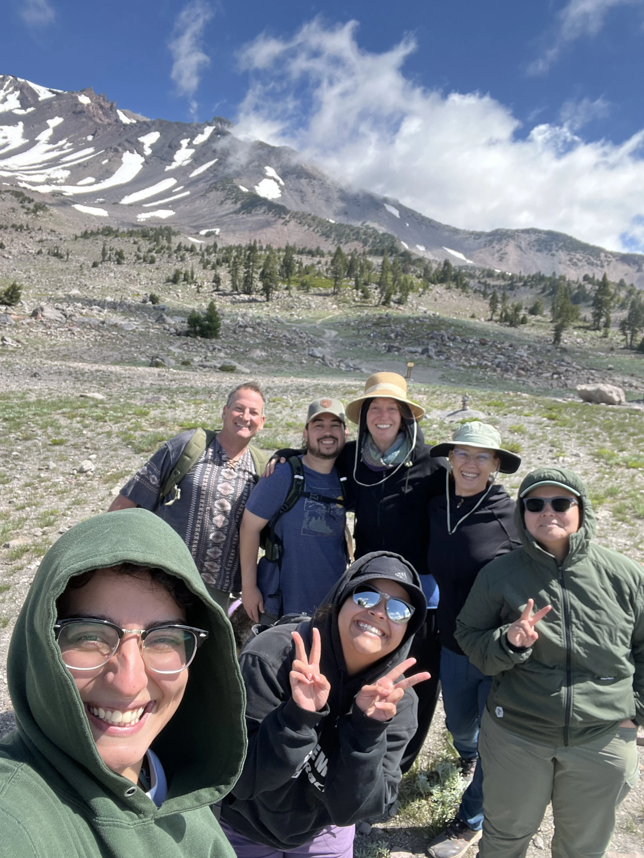 Group of six hikers in a mountainous landscape with snow-capped peaks, some trees, and partly cloudy sky, smiling and posing for a photo.