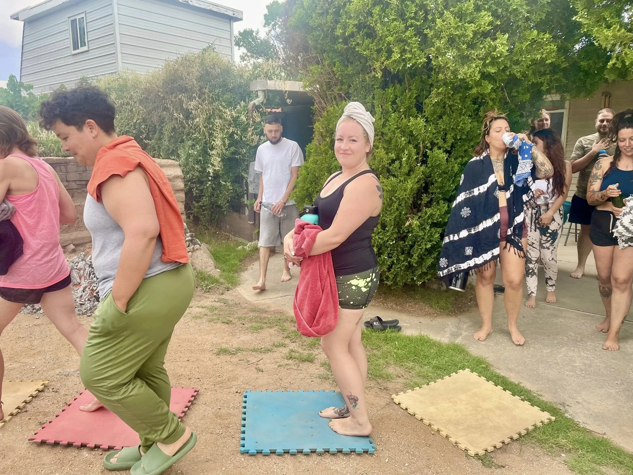 People gathered outdoors during a casual social event, some standing on foam mats on the ground, with a house and greenery in the background. Some are dressed in casual summer clothing, and one woman in the foreground is barefoot, holding a towel and smiling at the camera.
