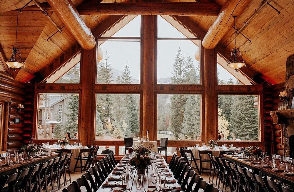 Rustic log cabin wedding reception venue with long decorated tables, set dinnerware, floral centerpieces, and mountain views through large windows