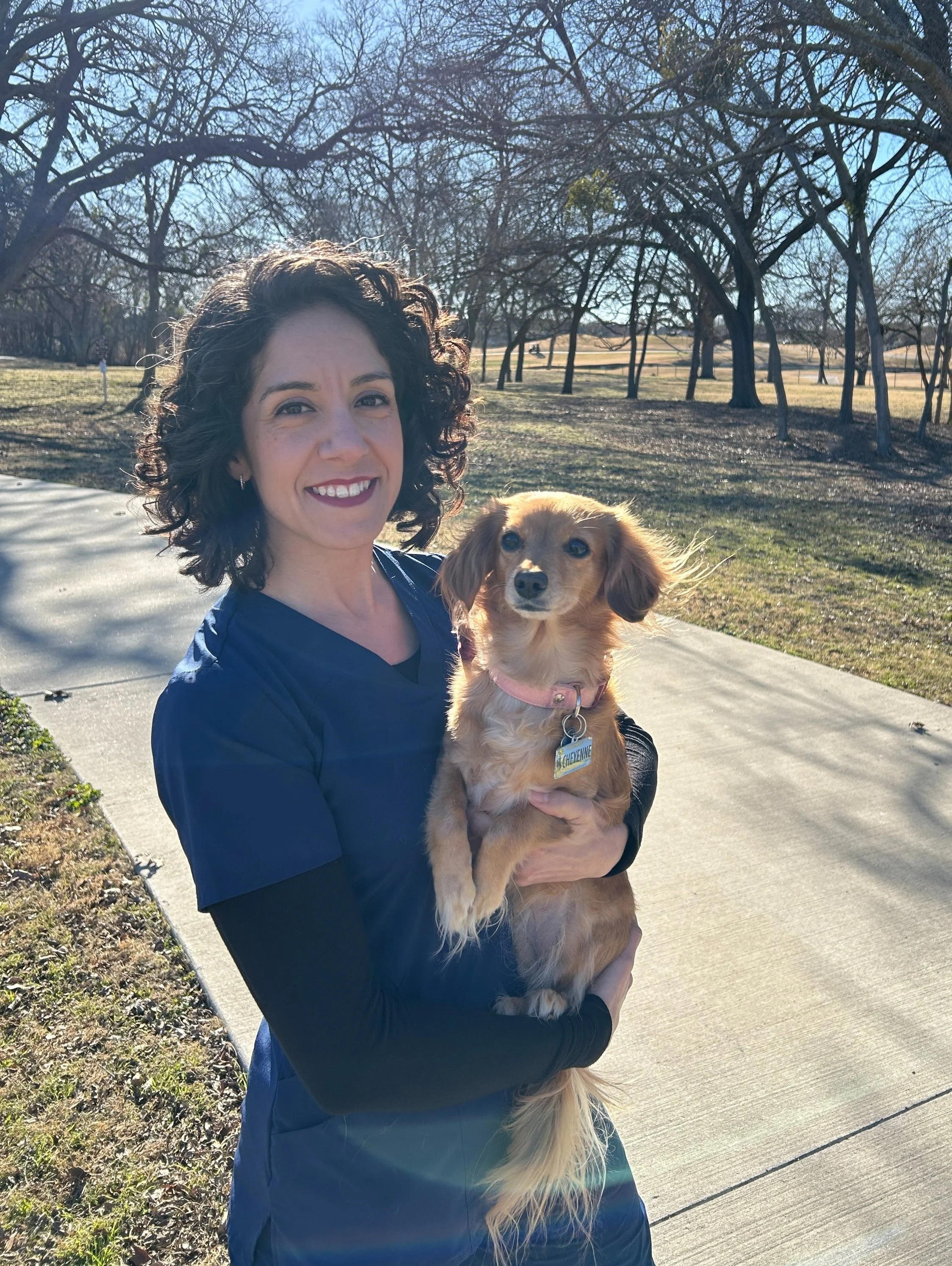 A woman with curly hair smiling and holding a small long-haired dog with a pink collar in a park on a sunny day.