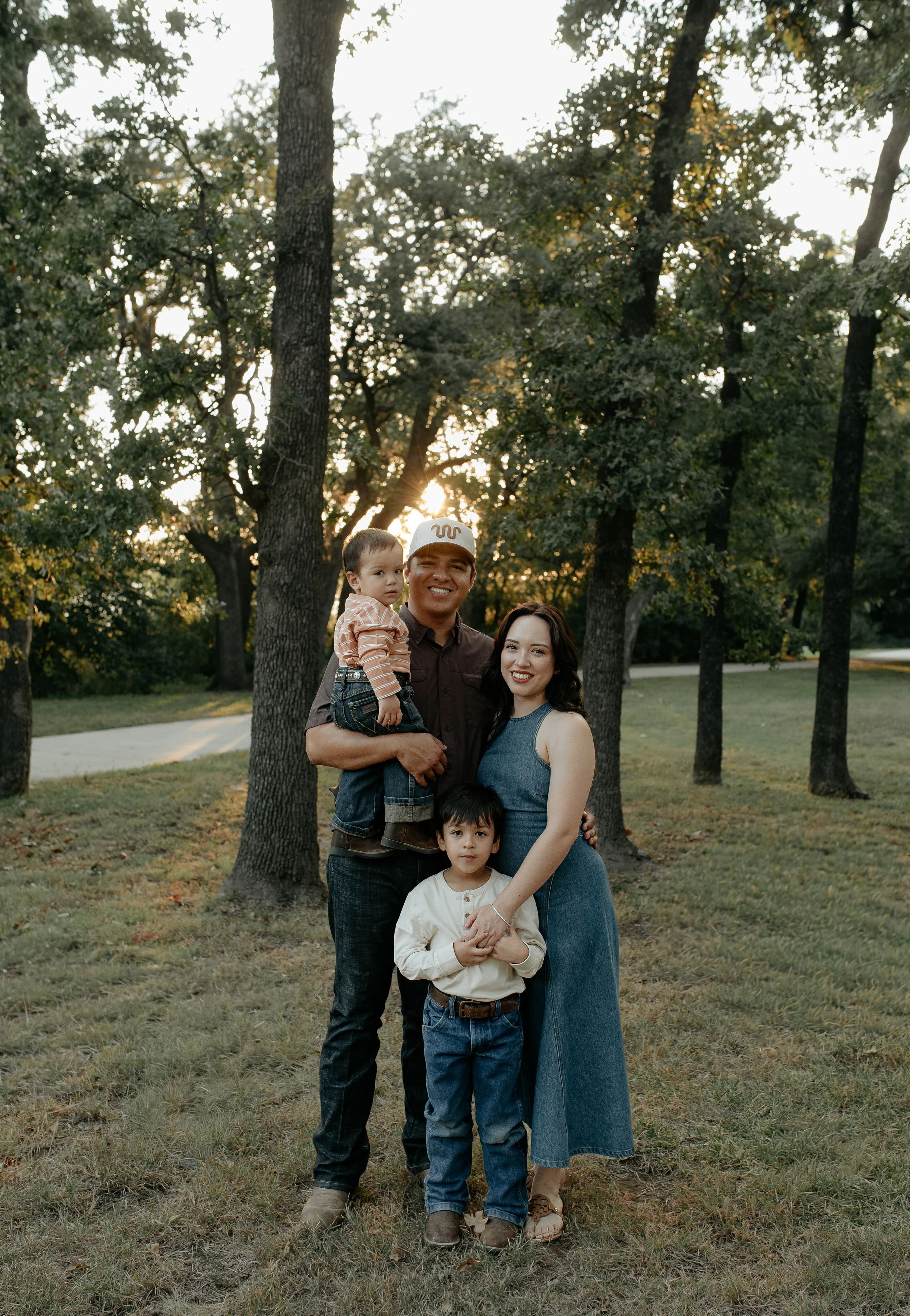 A smiling family of four standing outdoors in a grassy park with trees and sunset in the background.