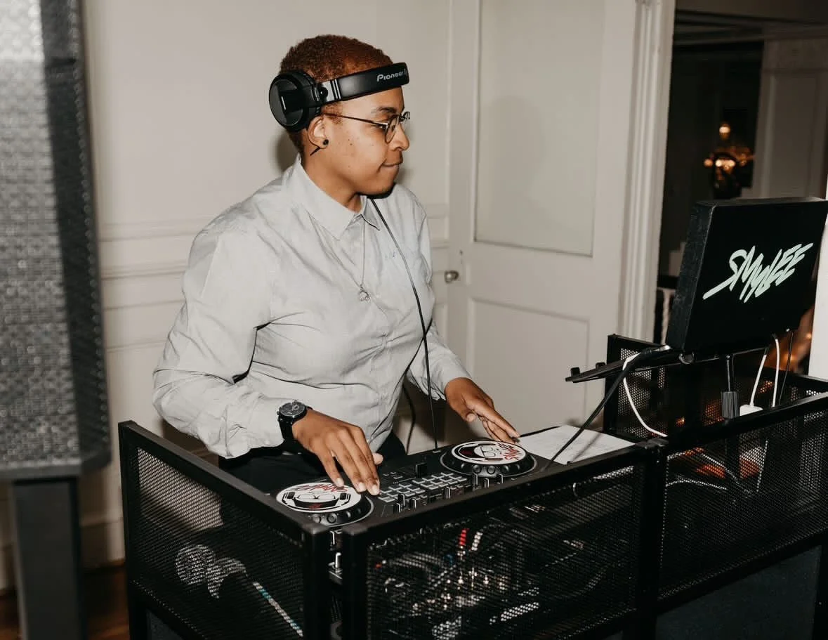 A woman DJ wearing headphones and glasses, standing behind a DJ controller with a laptop, indoors.