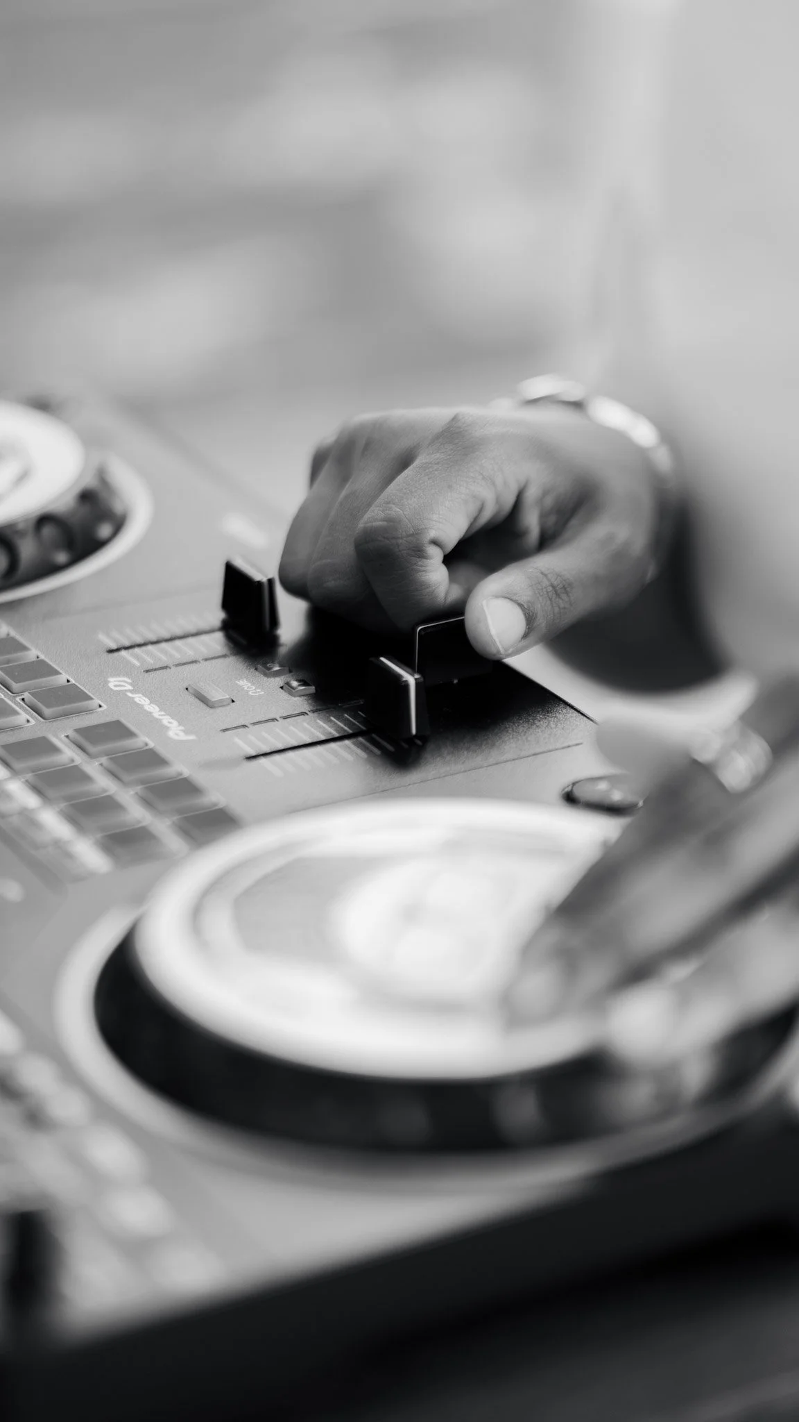 Close-up of a hand adjusting controls on a DJ mixing console, with turntables in the foreground, in black and white.