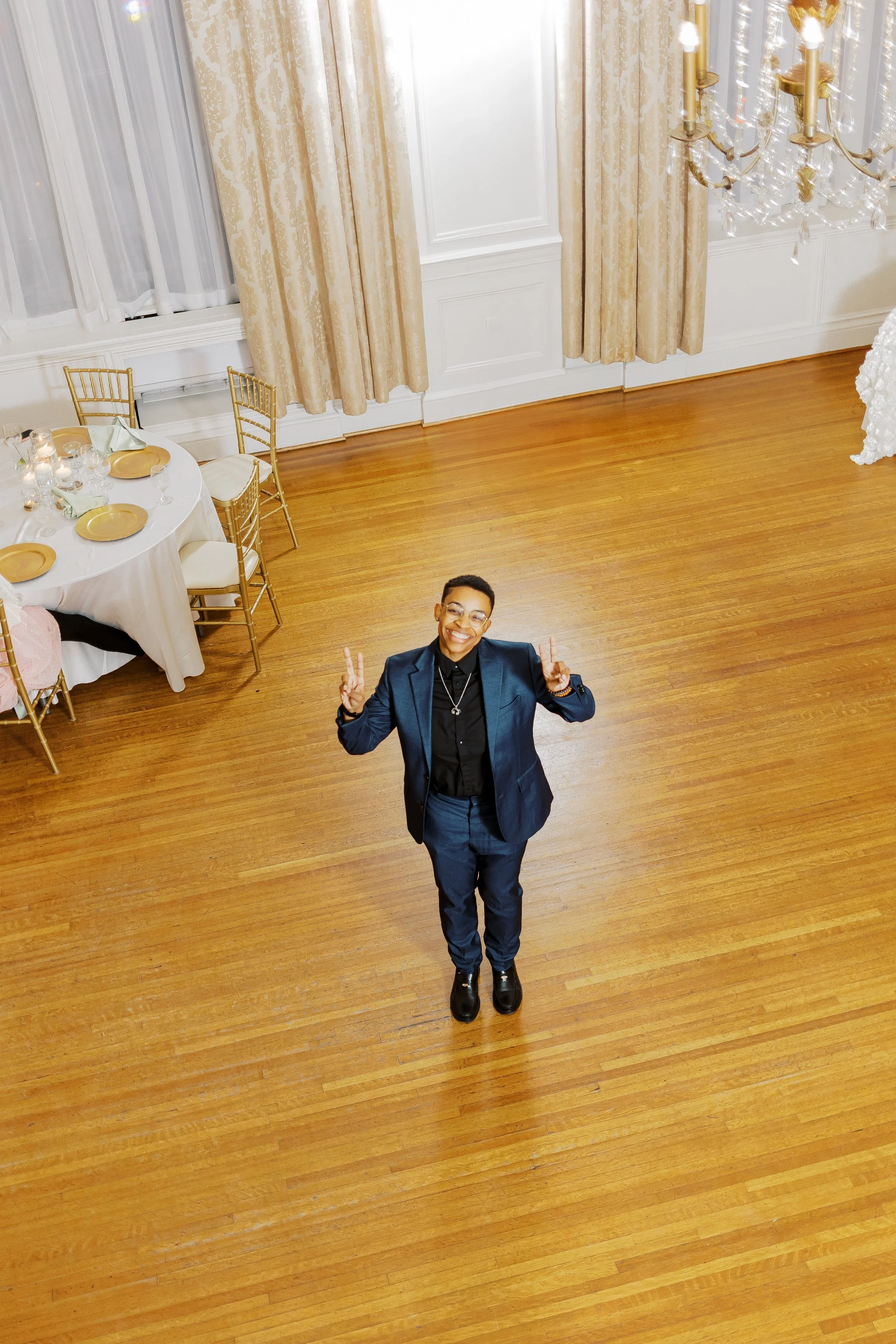 A person standing in a large, elegantly decorated room with wooden floors, dressed in a dark blue suit and black shoes, smiling and making peace signs with both hands. The room has a chandelier, beige curtains, and a table set with gold plates and ca