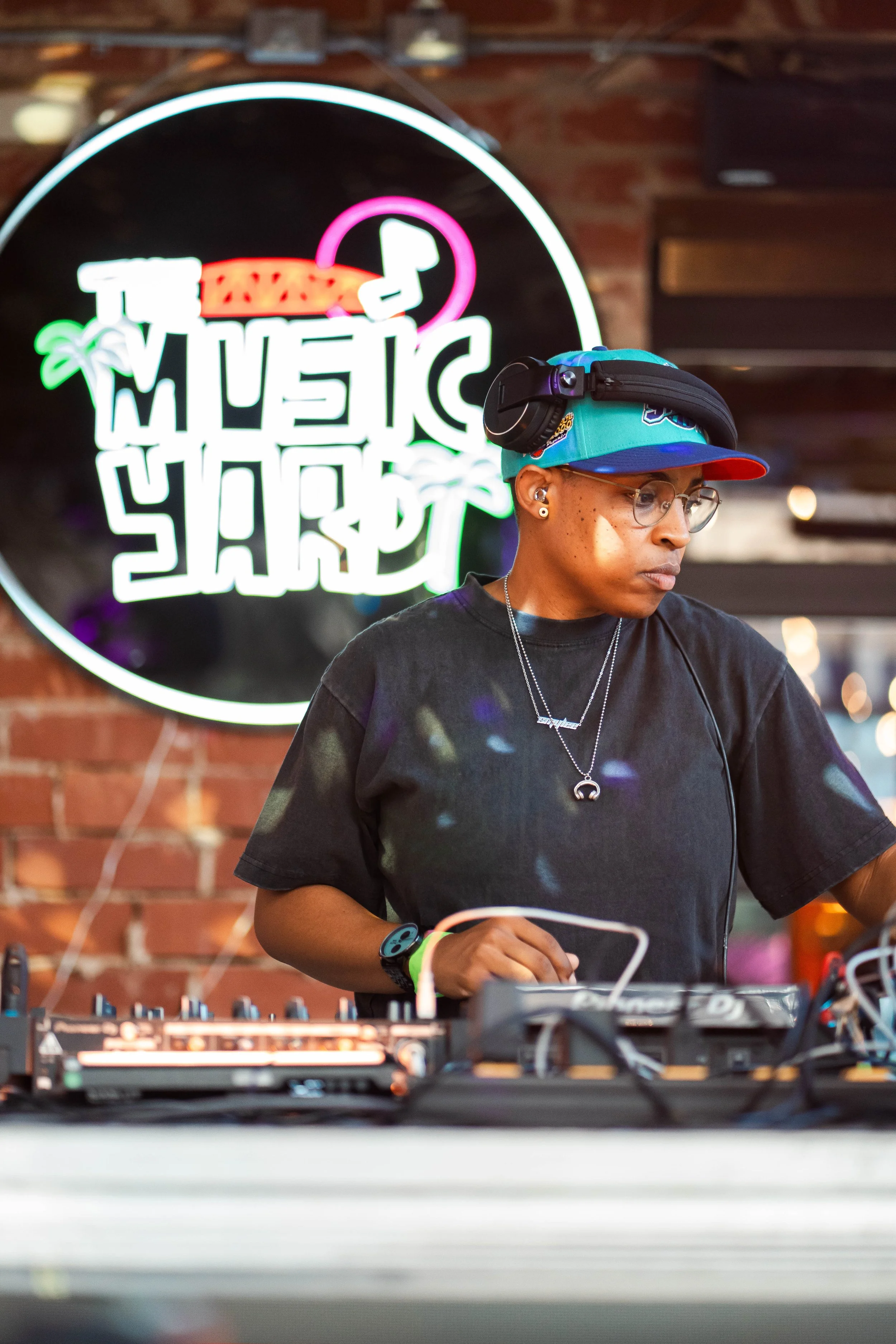 DJ Smylee is standing behind a DJ booth with a neon lit sign behind her saying "The Music Yard"