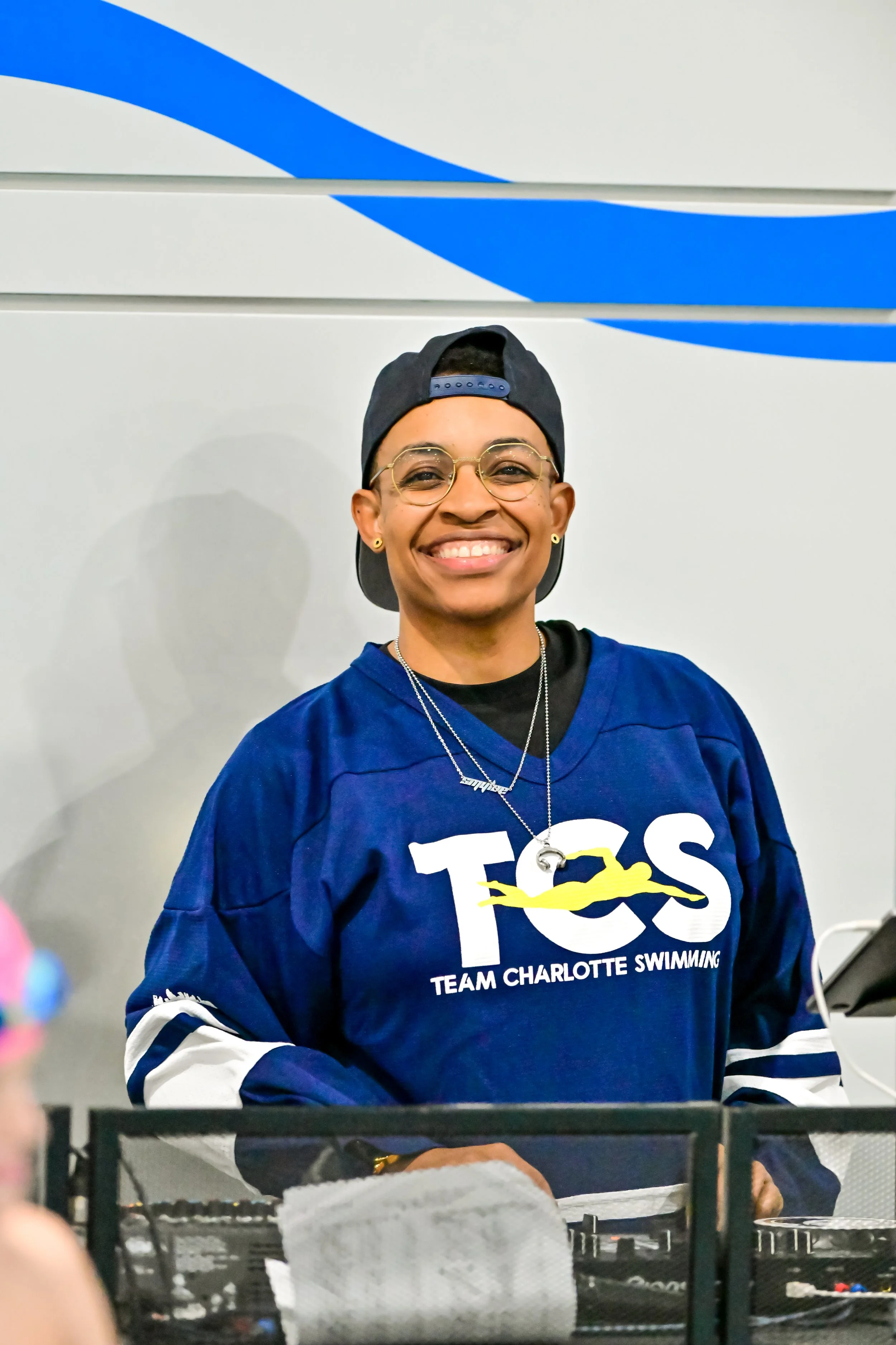 Young woman wearing glasses, a backwards cap, and a blue Team Charlotte Swimming jersey, smiling at the camera.